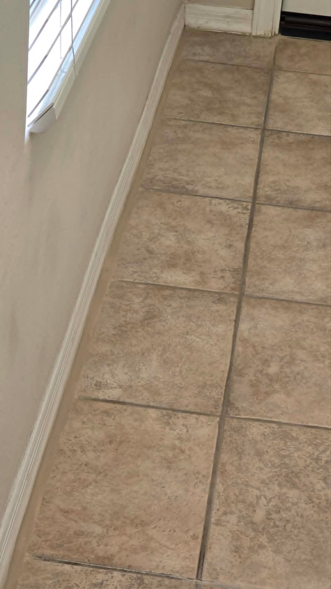 Ceramic tile floor in a Chandler AZ home with a cracked, eroded perimeter grout joint pulling away from the baseboard, showing soil packed into the open joint before tile and grout repair